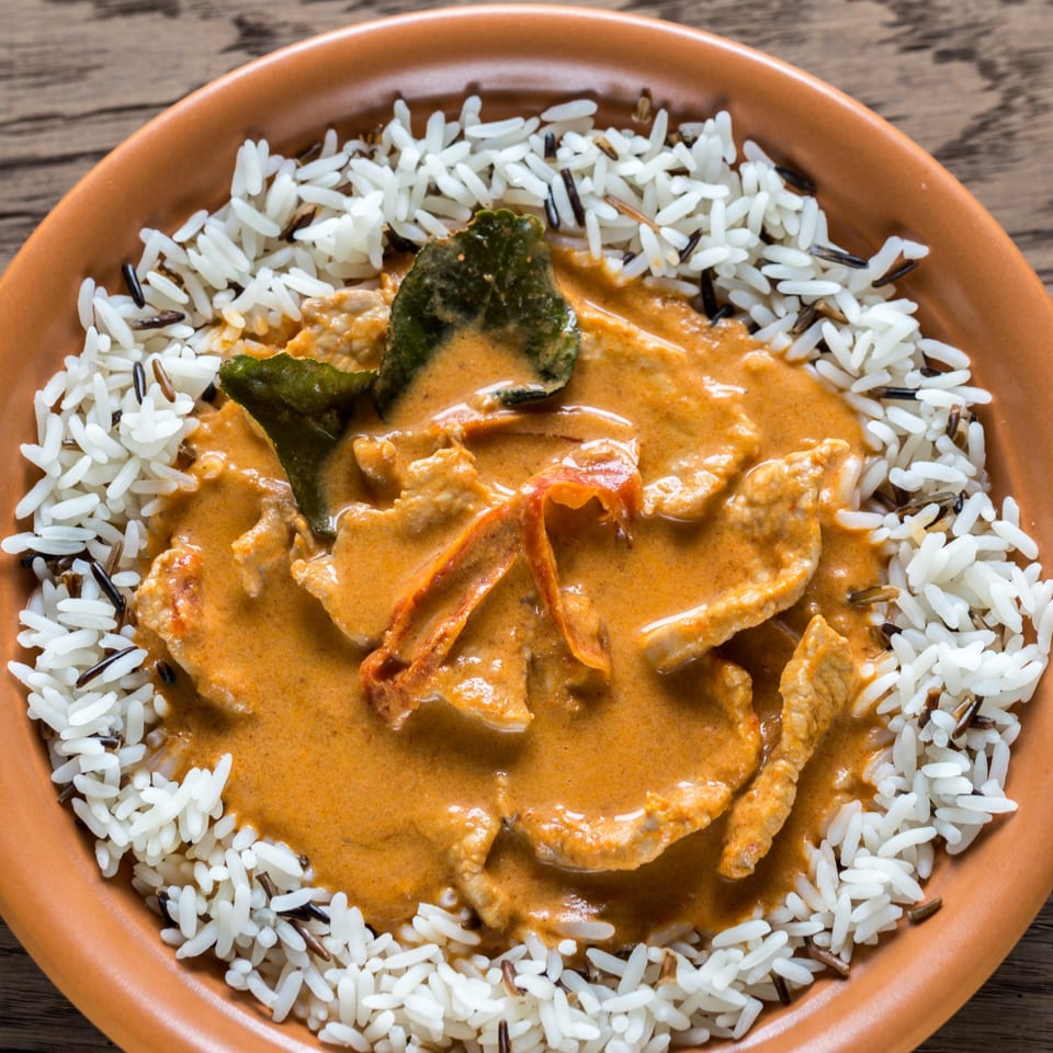 Close-up of a vibrant red Thai curry with meat and vegetables, served with white and wild rice.