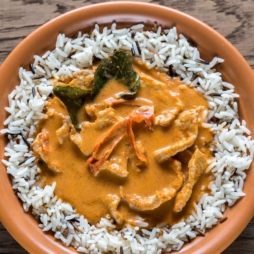 Close-up of a vibrant red Thai curry with meat and vegetables, served with white and wild rice.